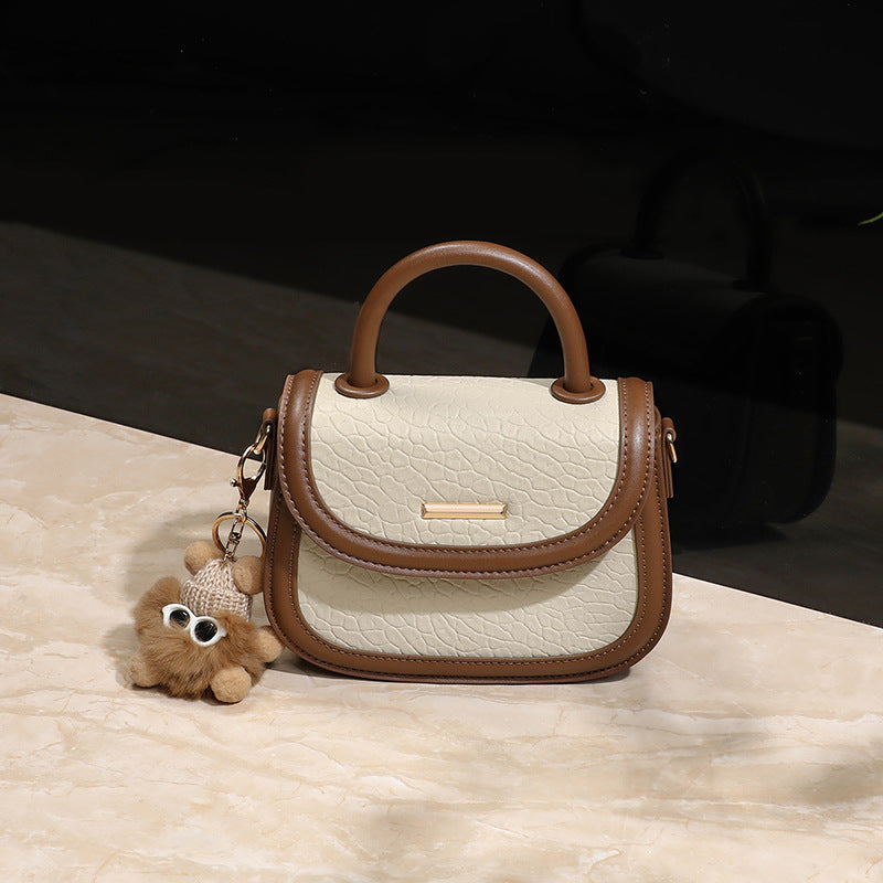 Small handbag with brown handle and trim, featuring a teddy bear keychain on a light surface.