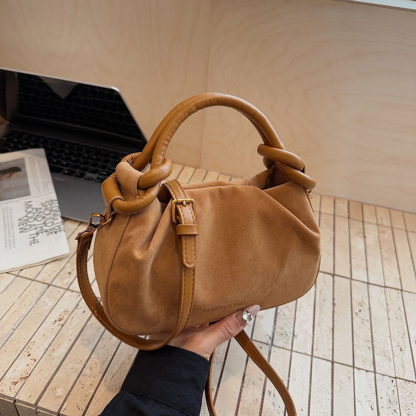 Brown handbag held by a person on a wooden floor with a laptop and magazine in the background.