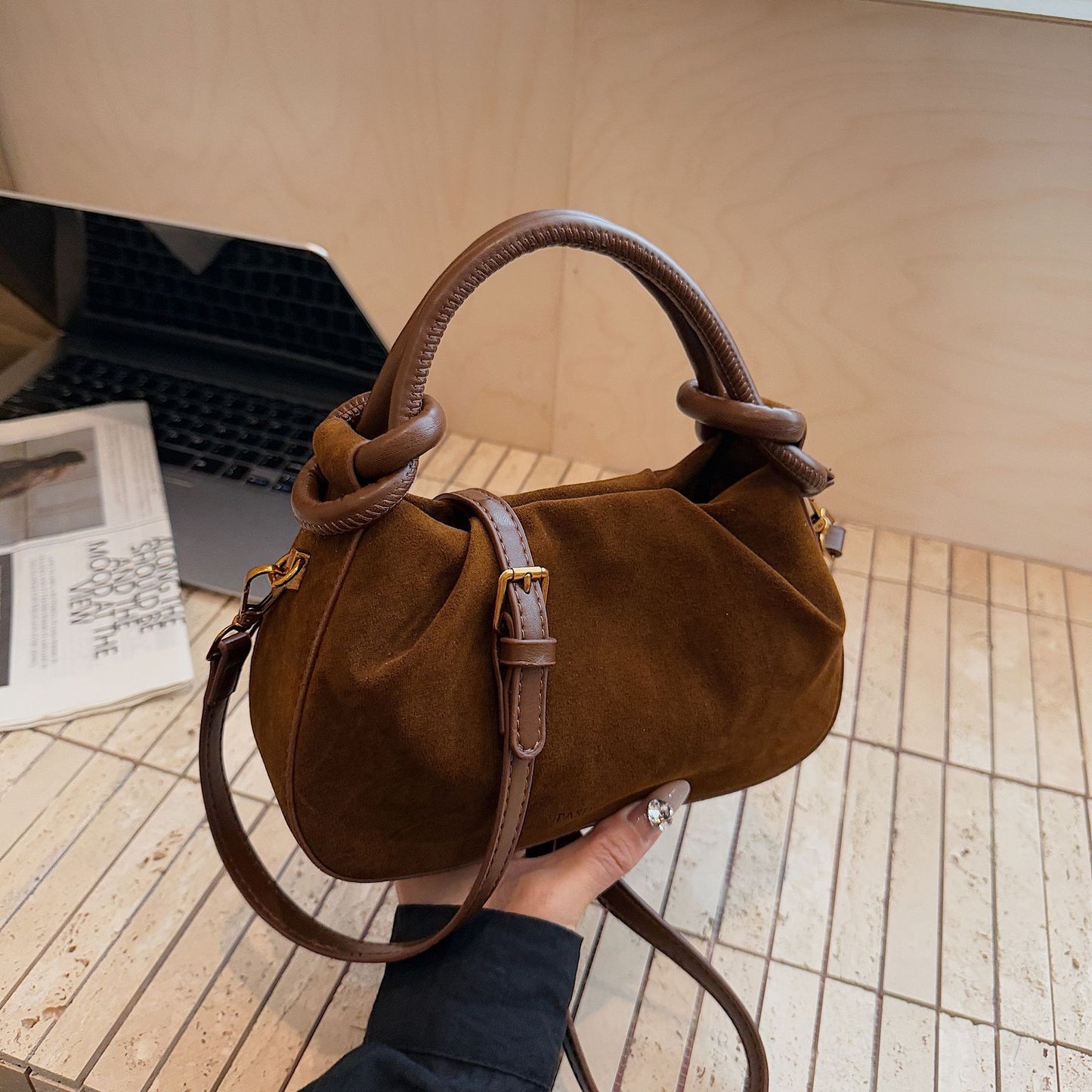 Brown handbag held by a person on a wooden floor with a laptop and magazine in the background.